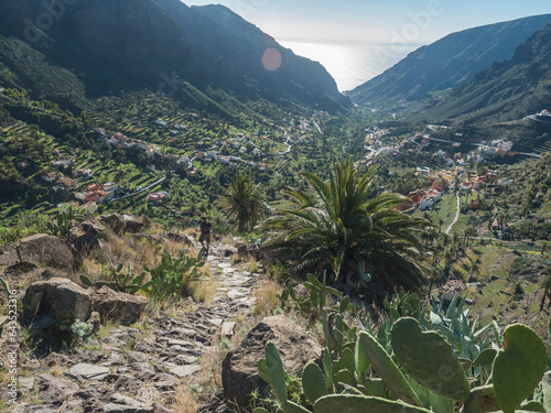 Man hiker on steep old stone road to El Retamal village. Green valley with palm trees, terraced fields and traditional colorful houses. Valle Gran Rey, La Gomera, Canary Islands, Spain, Europe.