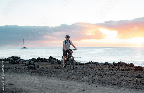 Happy active senior cyclist man at sea at sunset light with electric bicycle running on the beach - elderly man with helmet enjoying healthy lifestyle and freedom