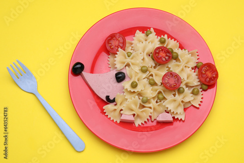 Creative serving for kids. Plate with cute hedgehog made of delicious pasta, sausages and tomatoes on yellow table, flat lay