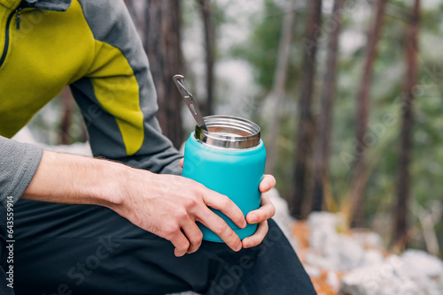 Embracing nature's flavors: A hiker enjoys a hot breakfast, holding a thermos amid a serene outdoor setting.
