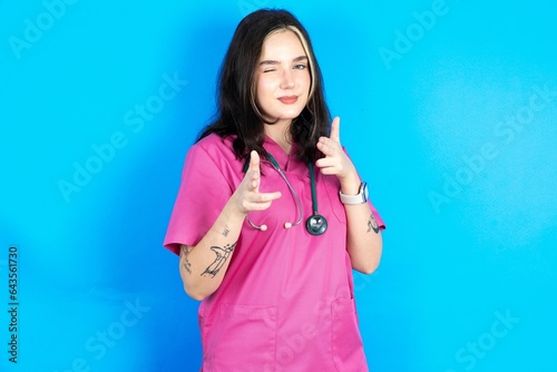 Joyful young beautiful doctor woman wink and points index fingers at camera, chooses someone, makes finger gun pistol. photo