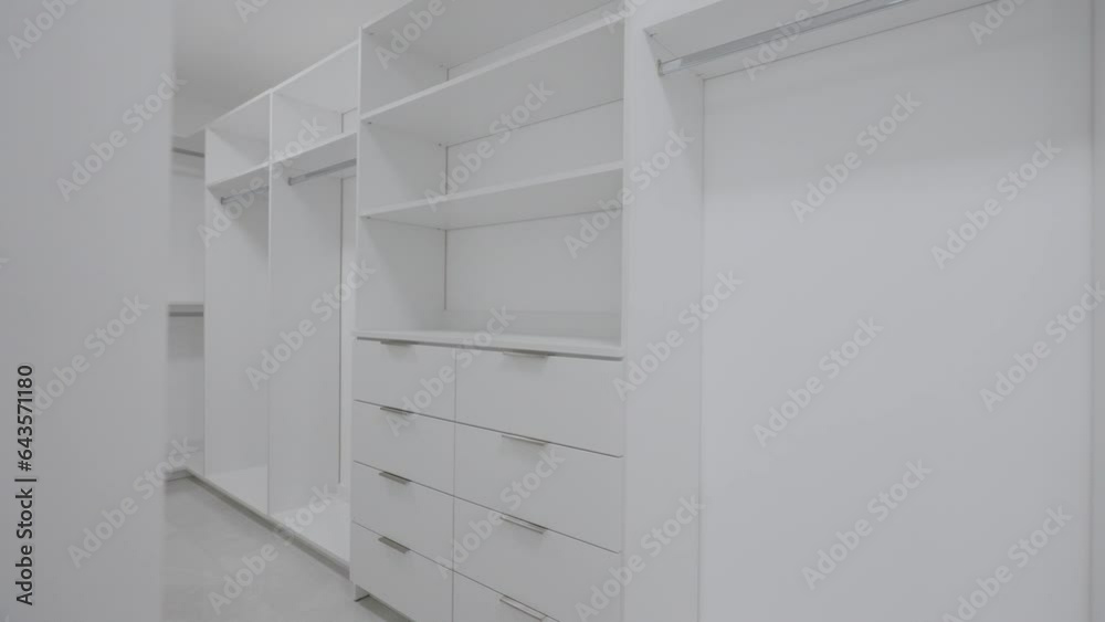 Large spacious empty walk-in closet with white cabinets, drawers, and holders for hangers. Beautiful wardrobe for placing clothes and shoes. Interior design concept