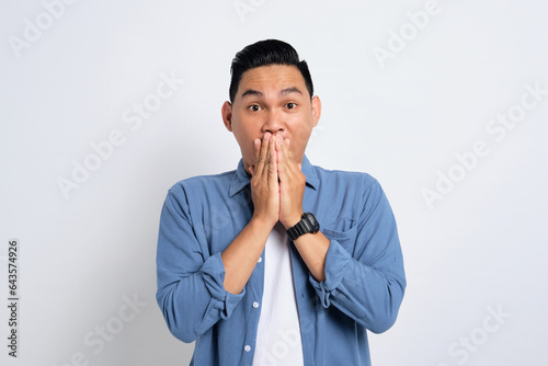 Surprised young Asian man in casual shirt covering mouth with hand, looking at camera with amazed facial expression isolated on white background