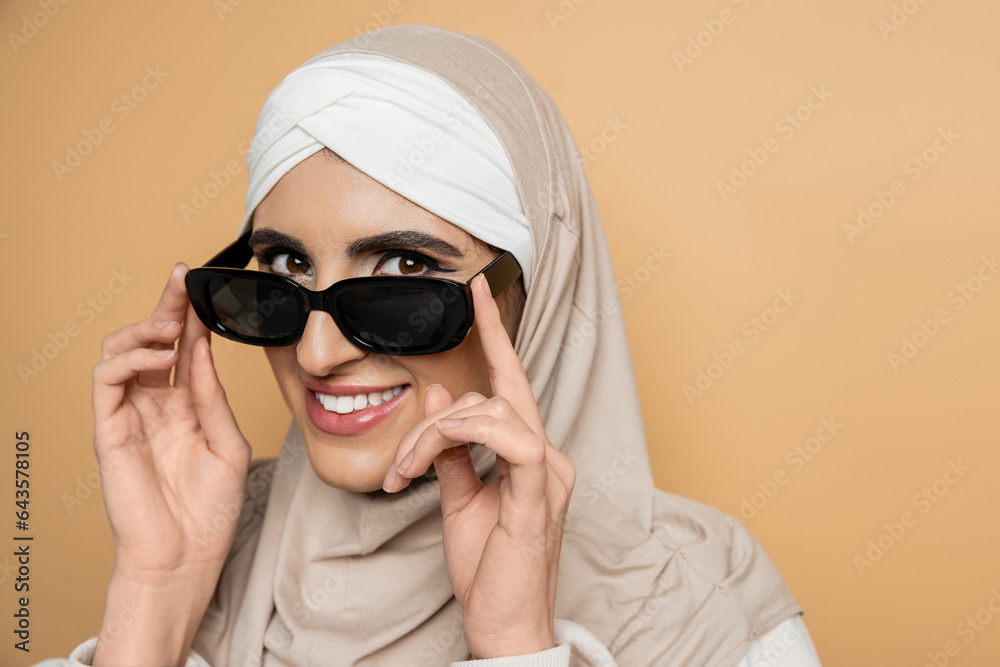 smiling muslim woman in hijab looking at camera over stylish sunglasses on beige, modern lifestyle smiling muslim woman in hijab looking at camera over stylish sunglasses on beige, modern lifestyle