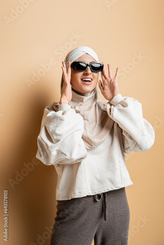 amazed muslim woman in hijab, sweatshirt and trendy casual attire adjusting sunglasses on beige