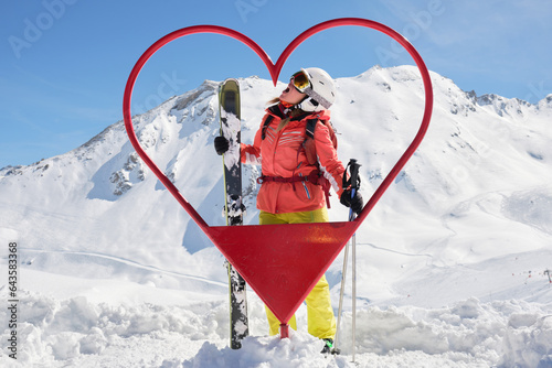 Woman skier poses inside a heart shaped frame at Tignes - Val d'Isere ski resort, France, with all mountan skis and red outfit, on a sunny day. Tourism, activity, Winter, snow. photo