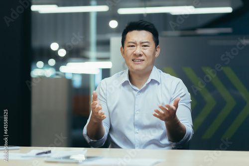 Angry and displeased young asian man businessman, office worker talking emotionally to camera on ringing while sitting at desk.