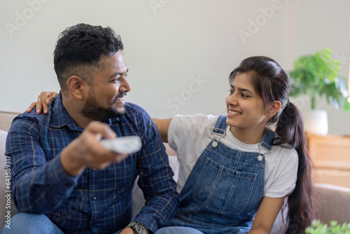 Happy beautiful young couple indian sitting on couch, embracing, watching TV together at home photo