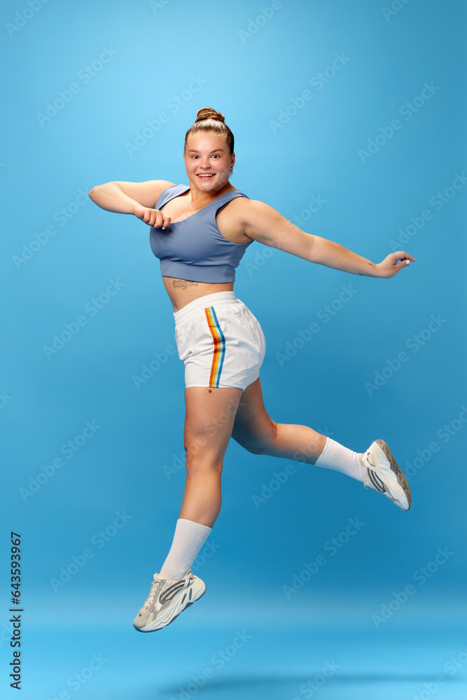 Full length portrait of young smiling fat overweight woman wearing sportswear doing exercise, jumping isolated on studio blue background.