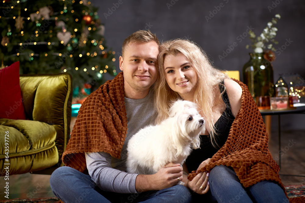 Lovely romantic couple with their dog near Christmas tree in festive aesthetic cozy home interior. Candid, true moment. Good mood and having fun together. Christmas love story Lovely romantic couple with their dog near Christmas tree in festive aesthetic cozy home interior. Candid, true moment. Good mood and having fun together. Christmas love story