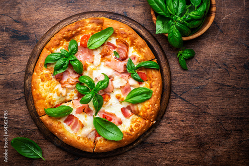 Hot pizza with ham, pancetta, mozzarella cheese, tomato sauce and green basil, rustic wooden table background, top view