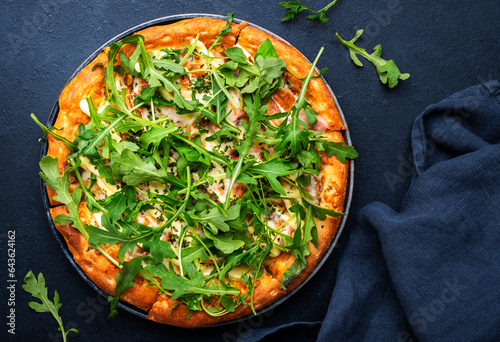 Hot pizza with mozzarella cheese, ham, pesto and fresh arugula, black table background, top view