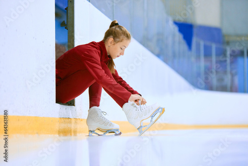 Картината върху платно Beautiful girl in red sportswear preparing for training session on ice rink arena, figure skater tying laces on skates