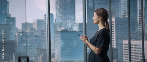 Caucasian Female Project Manager Standing in Modern Office Building Hallway With Big City View And Using Smartphone. Professional Woman Reading Emails, Checking Tasks, Texting Online. Anamorphic Shot photo