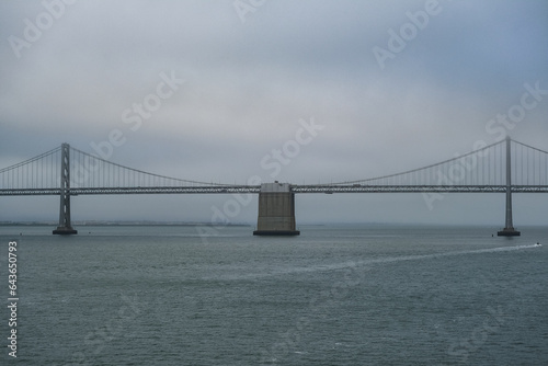 Grey metal steel suspension Bay bridge in San Francisco Bay Area with coast and city downtown skyline silhouette on cloudy day from outdoor cruiseship cruise ship deck