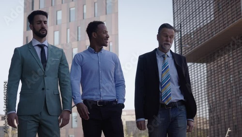 Three diverse business men communicating in free time walking outdoors. Multicultural suit coworkers discussing contract strolling together to office. Professional people and positive labor relations  photo