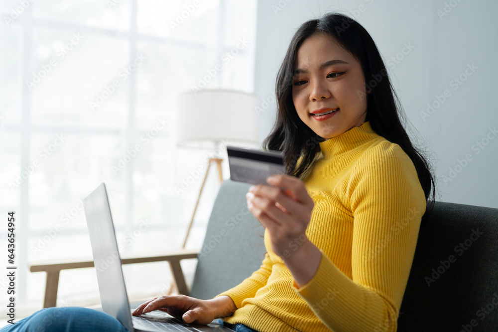 Young Asian woman holding credit card and smartphone sitting on sofa at home doing online banking transactions in home online shopping in living room. online payment and ordering concept. Young Asian woman holding credit card and smartphone sitting on sofa at home doing online banking transactions in home online shopping in living room. online payment and ordering concept.
