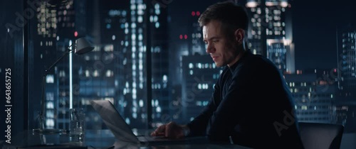 Successful Handsome Businessman Working on Laptop Computer in Big City Office Late At Night. Financial Investment Analyst Checking Data And Works On Risk Management In Hedge Fund. Anamorphic Shot photo