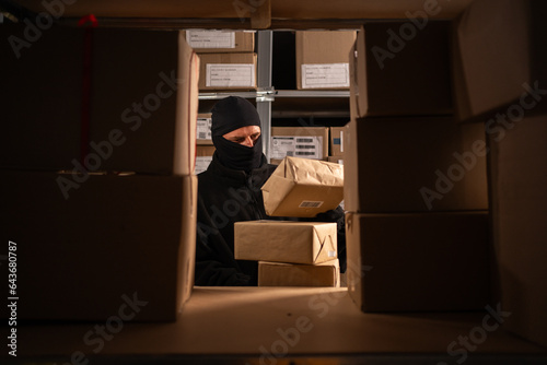 Fotografie Thief in black balaclava steals with a parcel in a warehouse at the night