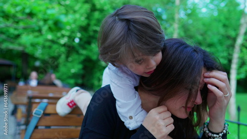 Abrasive little boy climbing on top of mother's neck harshly, showing little concern to mom's emotion in open air outside. parent in discomfort, parenting concept