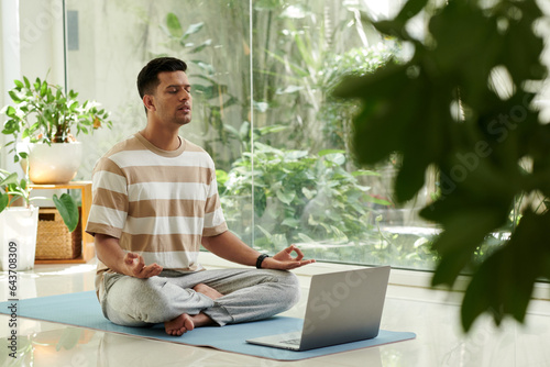 Fototapeta Naklejka Na Ścianę i Meble -  Young serene man in activewear keeping eyes closed during home yoga practice while sitting on mat in pose of lotus in front of laptop screen