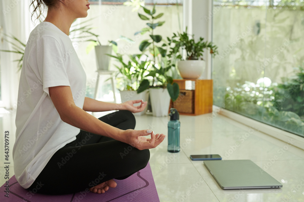 Cropped shot of young active woman in white t-shirt and black leggins sitting in pose of lotus on mat while practicing yoga exercises Cropped shot of young active woman in white t-shirt and black leggins sitting in pose of lotus on mat while practicing yoga exercises
