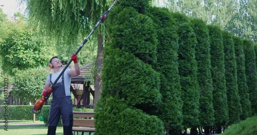 Strong experienced gardener trimming hedge with modern electric machine on backyard. Handsome caucasian man in overalls working with gardening tool outdoors. Concept of seasonal work. 