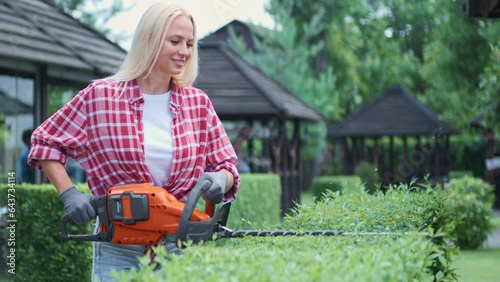 Smiling blonde girl in red plaid shirt holding trimmer for shaping bushes in garden. Front view of female gardener taking care of boxwood with electric lopper in spring time. Concept of seasonal work. photo