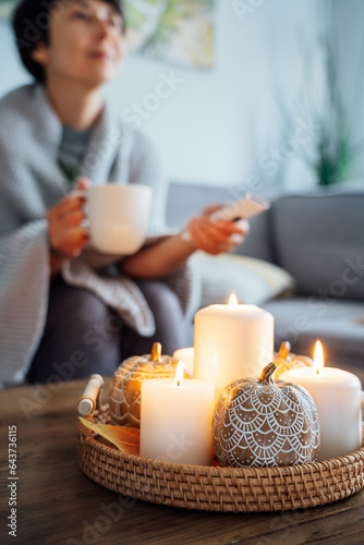 Focus on autumn fall cozy mood decor composition with a blurred woman covered plaid selecting movie with remote controller and drinking tea on the sofa. Movie night party at home. Cozy relaxing time.