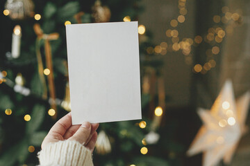 Christmas card mock up. Hand holding empty greeting card on background of stylish decorated christmas tree with golden lights. Space for text. Season greetings template and vintage ornaments