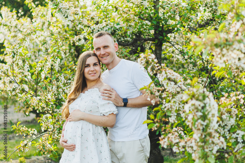 Tender romantic pregnant couple hugging in blooming apple spring garden. Expectant parents in the park of white flowering trees. A couple is expecting a baby Tender romantic pregnant couple hugging in blooming apple spring garden. Expectant parents in the park of white flowering trees. A couple is expecting a baby