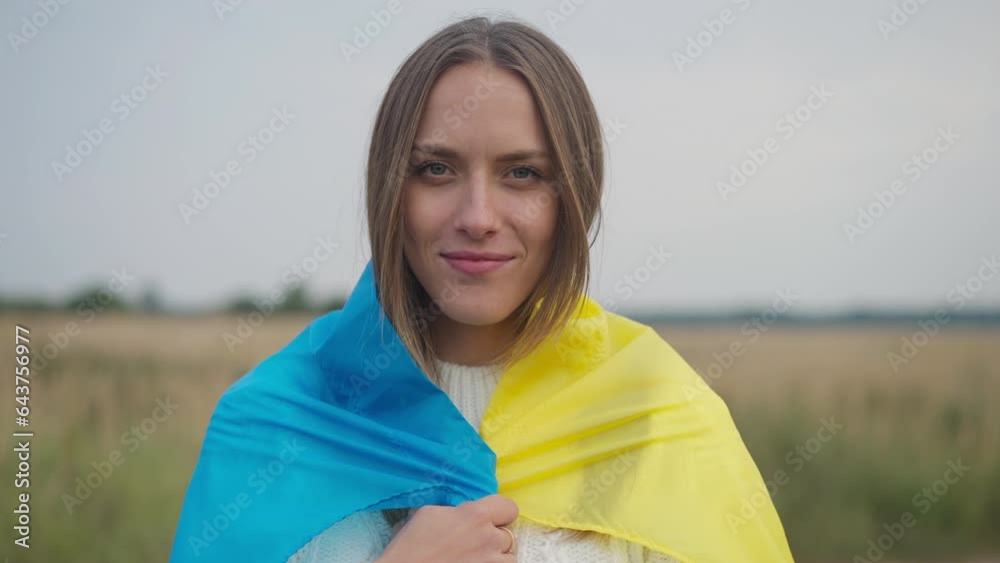 Front view confident woman putting Ukrainian flag on shoulders smiling looking at camera. Portrait of charming attractive young Caucasian lady posing in wheat field on cloudy autumn day outdoors Front view confident woman putting Ukrainian flag on shoulders smiling looking at camera. Portrait of charming attractive young Caucasian lady posing in wheat field on cloudy autumn day outdoors