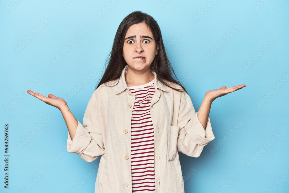 Young Caucasian woman on blue backdrop confused and doubtful shrugging shoulders to hold a copy space. Young Caucasian woman on blue backdrop confused and doubtful shrugging shoulders to hold a copy space.