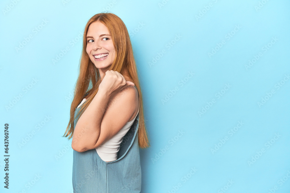 Redhead young woman on blue background points with thumb finger away, laughing and carefree. Redhead young woman on blue background points with thumb finger away, laughing and carefree.