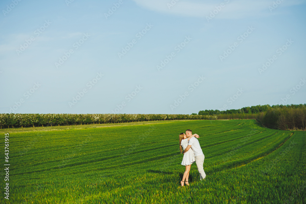 Pregnant couple hugging in a green spring field. Expectant parents in the park of white flowering trees. A romantic couple expecting a baby. A walk through a green field Pregnant couple hugging in a green spring field. Expectant parents in the park of white flowering trees. A romantic couple expecting a baby. A walk through a green field