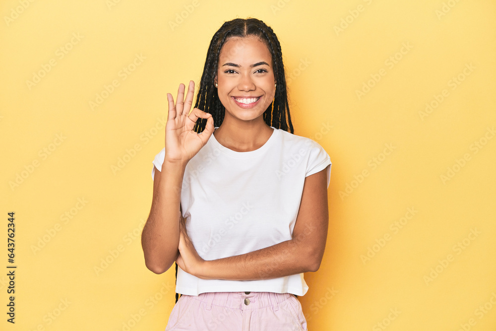 Young Indonesian woman on yellow studio backdrop winks an eye and holds an okay gesture with hand.