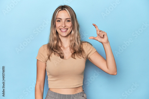 Obraz na płótnie Blonde young caucasian woman in blue studio holding something little with forefingers, smiling and confident