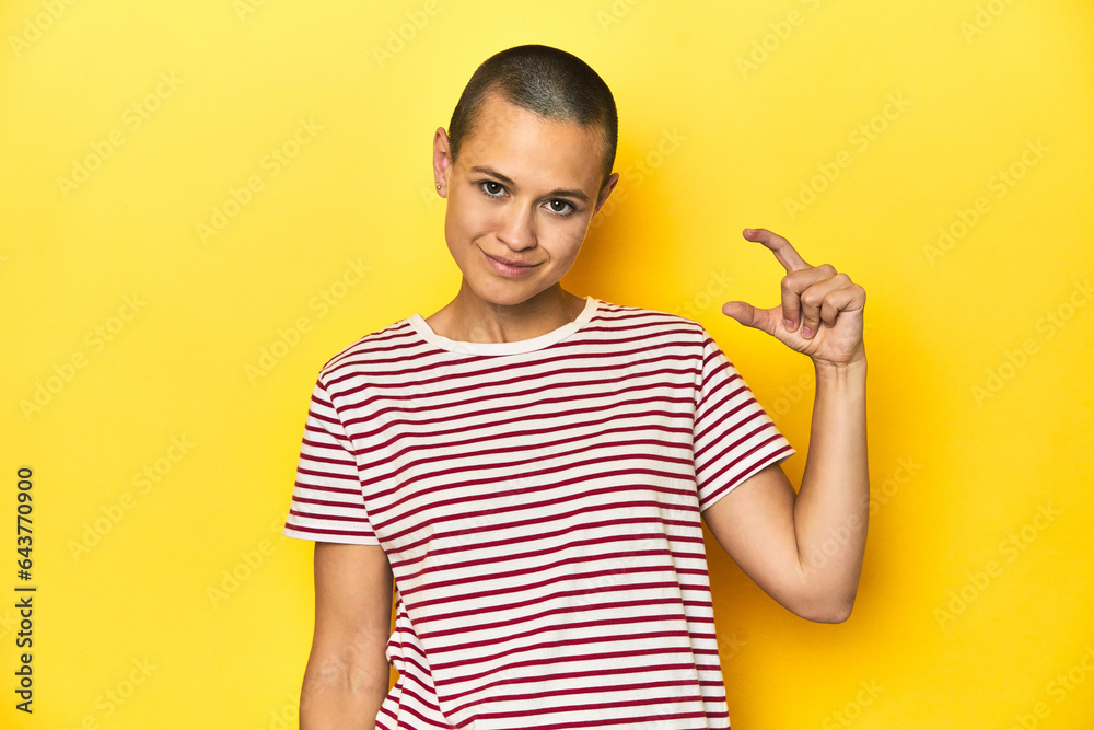 Shaved head woman in red striped tee, yellow backdrop holding something little with forefingers, smiling and confident. Shaved head woman in red striped tee, yellow backdrop holding something little with forefingers, smiling and confident.