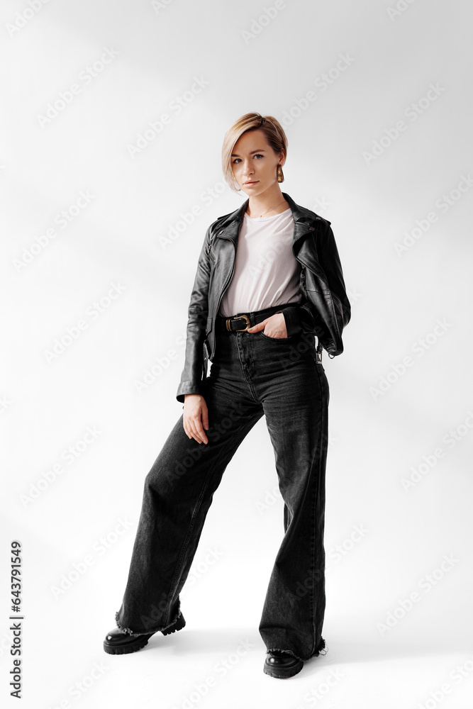 Full body of young woman with short blonde hair shows off her confident and stylish attitude in a black leather jacket and wide legged jeans, looking at camera against white background Full body of young woman with short blonde hair shows off her confident and stylish attitude in a black leather jacket and wide legged jeans, looking at camera against white background
