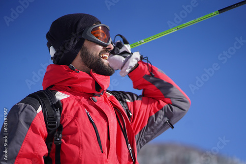 Cheerful man wearing ski equipment, smiling and holding a ski pole on a clear sunny weather outdoors. Male person with sport spirit outdoors.