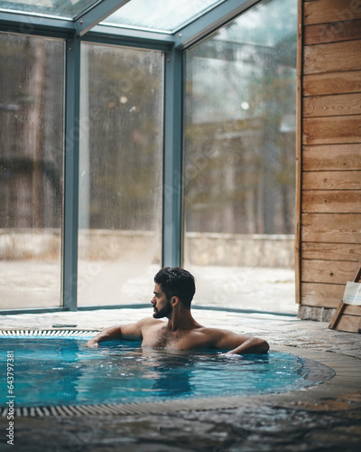 Handsome serious man with dark hair and beard sitting in the bathtub during the cold winter day. Good looking man relaxing in spa center.