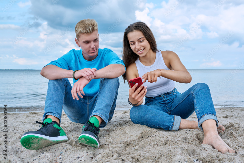 Teenage friends guy and girl sitting together on beach, talking looking at smartphone screen