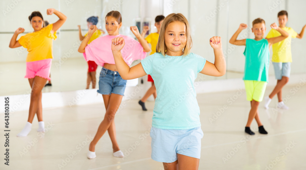 Fototapeta premium Portrait of cheerful preteen girl learning energetic dance moves with group of children in choreography class .