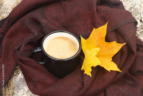 Hot milk coffee in enamel black mug wrapped in brown scarf with two yellow maple leaves as decor. Autumn hygge mood. Outdoors, picnic, camping, travel, trekking concept.