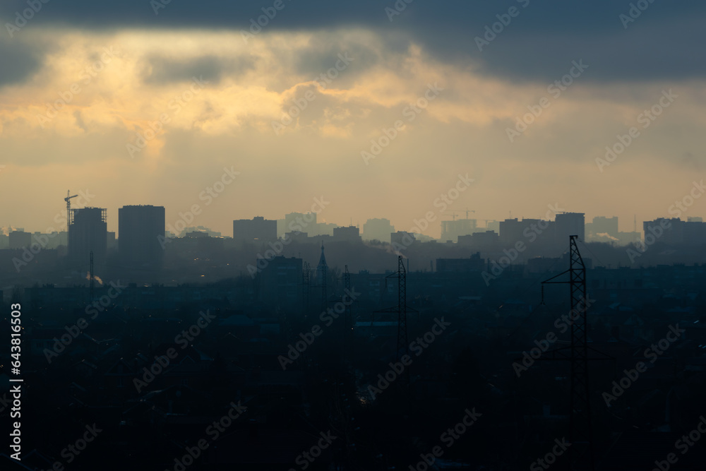 The rays of the setting sun break through the clouds, illuminating buildings in the haze of the city plunging into twilight