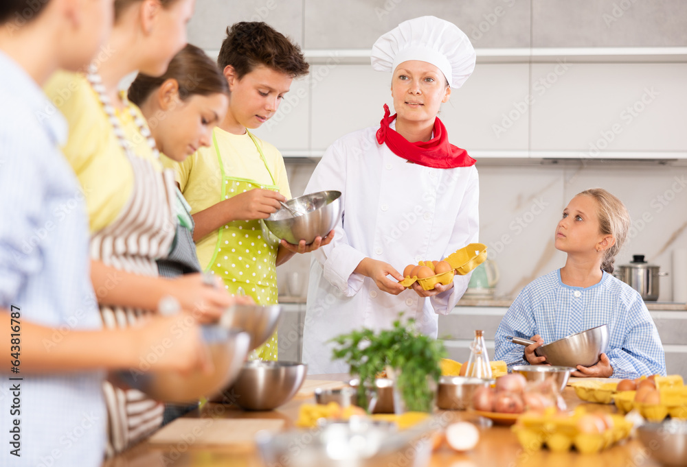 At culinary master class, experienced female chef in white uniform with egg tray in hands explains subtleties of dessert preparation to children. Woman shows sequence of actions during cooking