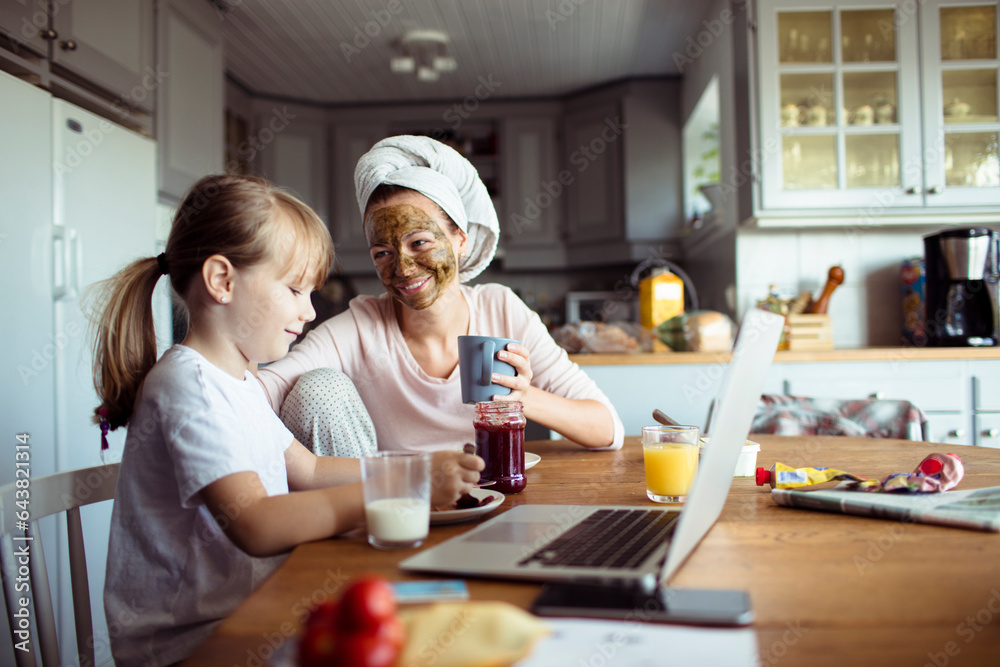 Young mother and daughter having breakfast and using a laptop in the morning in the kitchen Young mother and daughter having breakfast and using a laptop in the morning in the kitchen