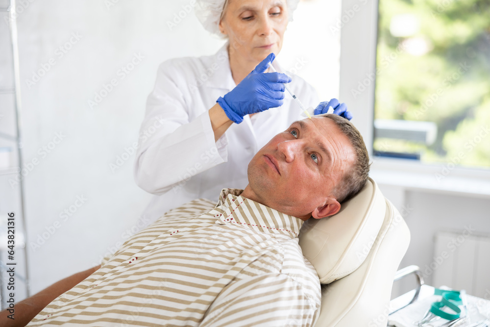 Obraz premium Adult man undergoing botulinum therapy session at aesthetic medicine clinic. Skilled woman cosmetologist making injections between eyebrows of patient with insulin syringe to correct facial wrinkles