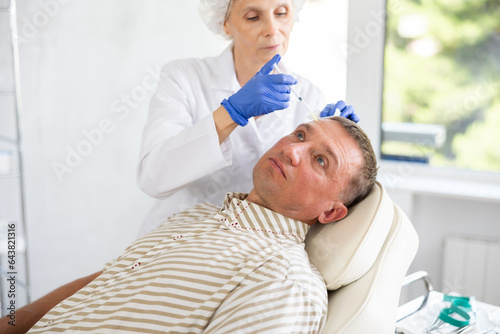 Adult man undergoing botulinum therapy session at aesthetic medicine clinic. Skilled woman cosmetologist making injections between eyebrows of patient with insulin syringe to correct facial wrinkles photo
