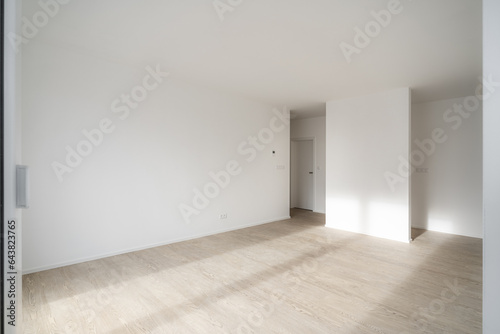 Empty unfurnished living room in a new apartment. Sunlight enters the room and hits the wooden floor. The walls are white and have electrical outlets  controls and other connections.